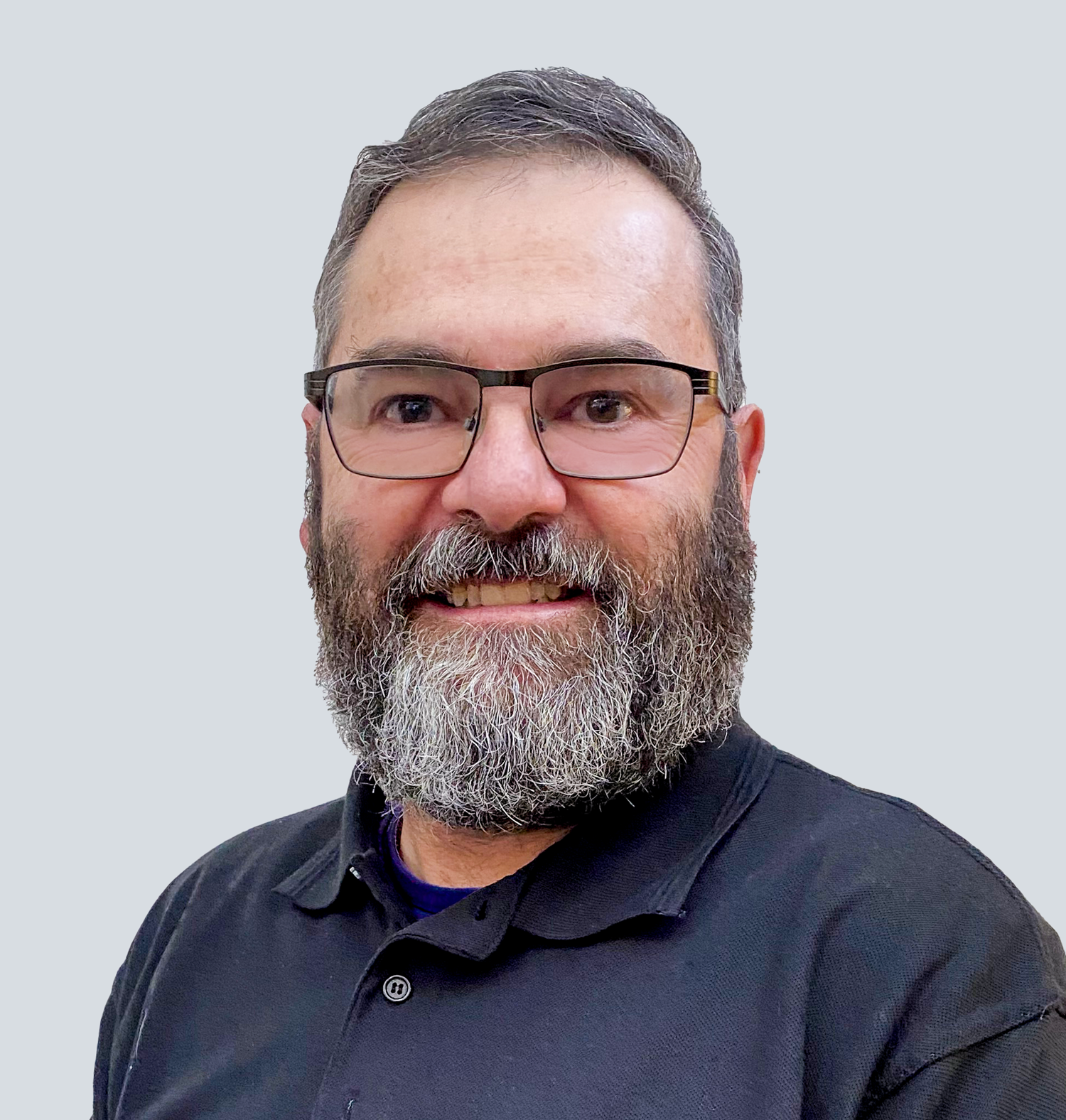 David, a Caucasian man with dark brown and grey hair and a short cropped dark brown and grey beard, wearing black framed glasses and a black polo shirt. He is standing in front of a light grey wall.